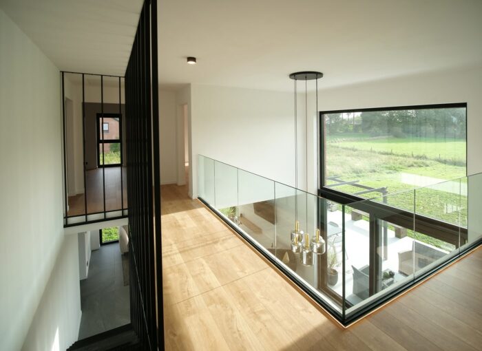 Vue intérieure moderne dune maison avec un plancher en bois et des balustrades en verre. Une grande fenêtre offre une vue sur un paysage verdoyant. Des plafonniers sont suspendus au-dessus et un escalier en métal noir est visible sur le côté.
