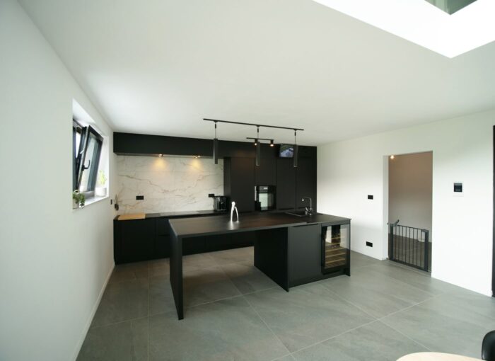Cuisine moderne avec comptoirs et armoires noirs, dosseret en marbre blanc et appareils électroménagers en acier inoxydable. Une cuisinière et un four encastrés se trouvent à gauche, avec un éclairage sur rail au plafond. La pièce a un design minimaliste avec une fenêtre à gauche.