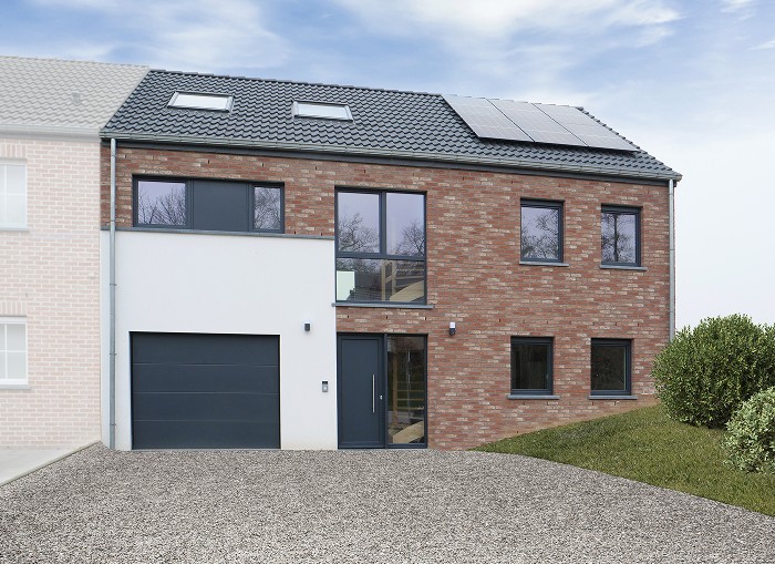 Une maison moderne en briques à deux étages avec de grandes fenêtres, une porte de garage grise et un toit en pente équipé de panneaux solaires. Une allée en gravier s'ouvre devant et un espace gazonné se trouve à droite. Le ciel est partiellement nuageux.