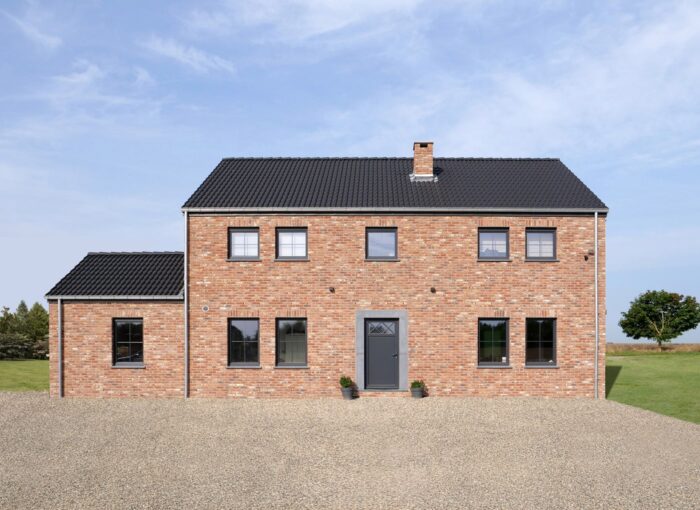 Une maison en briques de deux étages avec un toit en tuiles noires, six fenêtres à l'avant, une porte d'entrée grise et une petite annexe sur la gauche. La maison se trouve sur une allée de gravier avec de l'herbe et des arbres en arrière-plan sous un ciel bleu.