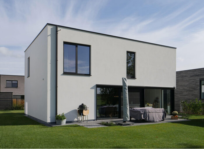 Modern huis met twee verdiepingen met een minimalistisch ontwerp, met grote ramen en een plat dak. Een kleine patio met een grill en overdekt meubilair is zichtbaar, omgeven door een goed onderhouden gazon. Andere soortgelijke huizen staan op de achtergrond.