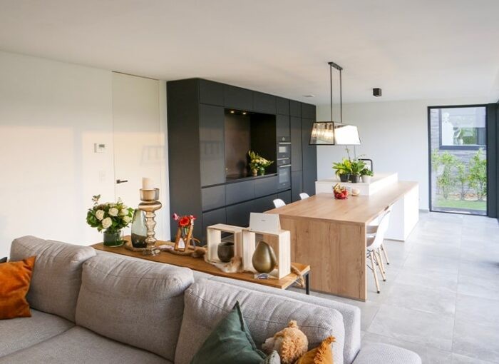 Moderne open keuken en woonkamer met een lichtgrijze bank. De keuken heeft een lang houten eiland, zwarte kasten en hanglampen. Een groot raam zorgt voor natuurlijk licht en decoratieve items en planten sieren de kamer.