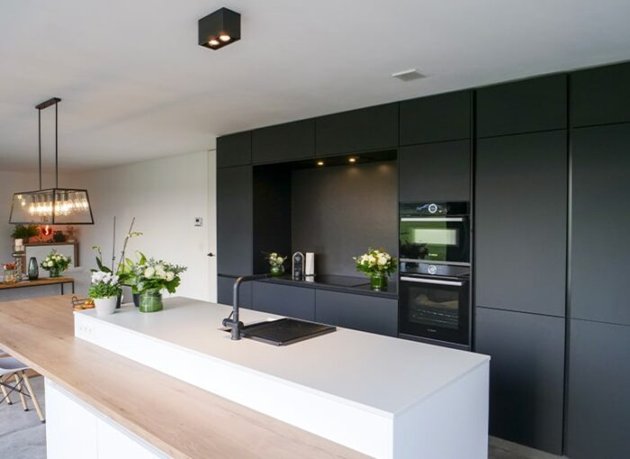 Moderne keuken met een wit kookeiland, zwarte kasten en ingebouwde oven. Het kookeiland heeft een gootsteen en inductiekookplaat. Er hangt een rechthoekige hanglamp boven en verschillende planten en bloemen voegen decoratieve accenten toe aan de hele kamer.