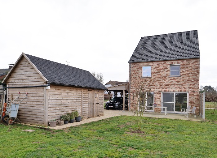 Bakstenen huis met twee verdiepingen en een donker dak, naast een houten schuur. Het huis heeft een open garage met een auto erin, een paar bomen en een grasveld. Er staan een kruiwagen en potplanten bij de schuur. De lucht is bewolkt.