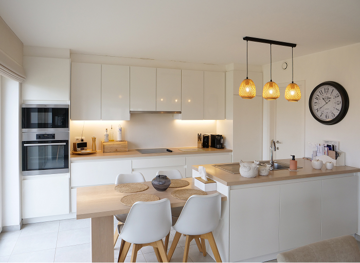 Moderne keuken met witte kasten, een houten eettafel en vier witte stoelen. Drie hanglampen hangen boven de tafel. Een wandklok en diverse keukenapparatuur zijn aanwezig. De ruimte is goed verlicht en georganiseerd.
