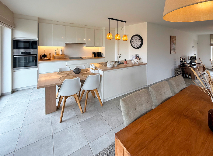 Moderne open keuken en eetkamer met witte kasten, een houten eettafel en stoelen. Hanglampen hangen boven een aanrecht met twee krukken. Een grote klok siert de muur en natuurlijk licht vult de ruimte via grote ramen.