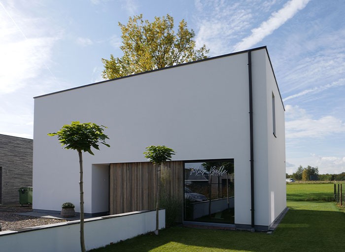 Modern wit, kubusvormig huis met grote ramen, minimalistisch design en strakke lijnen. Twee kleine bomen staan in de voortuin, met een groen veld en een helderblauwe lucht op de achtergrond.