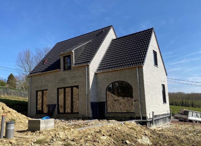 Een nieuw gebouwd huis met twee verdiepingen, lichte bakstenen muren, een zwart pannendak en grote ramen, omgeven door aarde en bouwmaterialen onder een helderblauwe lucht.