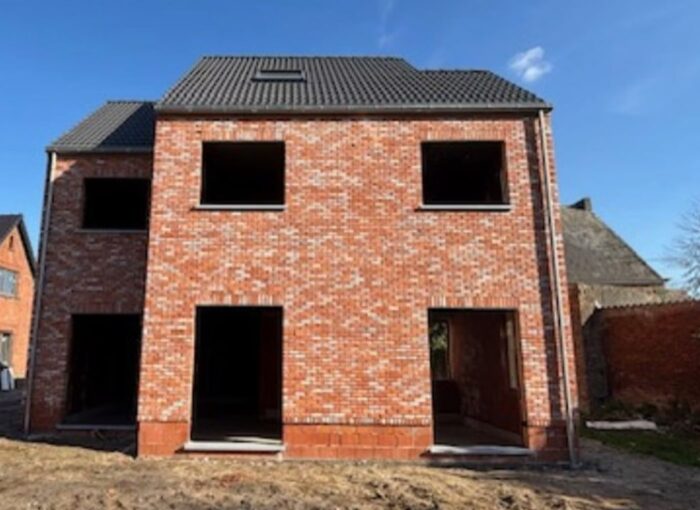 Een nieuw gebouwd huis van twee verdiepingen, van rode baksteen, met grote open ramen en deuren. Er zijn nog geen glas of deuren geplaatst en het dak is donker betegeld onder een helderblauwe lucht.