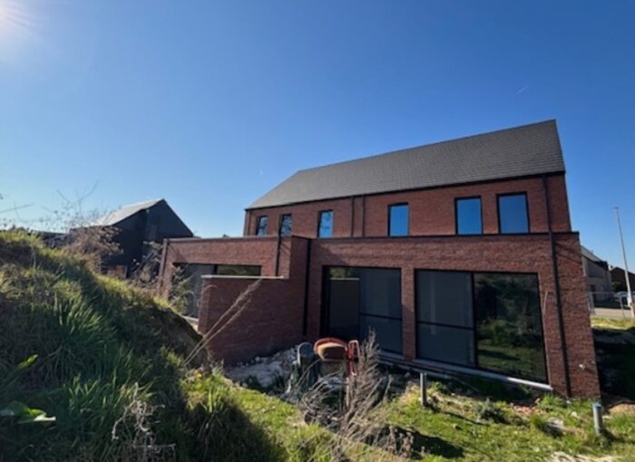 Een modern bakstenen huis met twee verdiepingen en grote ramen staat op een met gras begroeide helling onder een strakblauwe hemel. Bouwmaterialen en -gereedschappen liggen zichtbaar in de tuin.