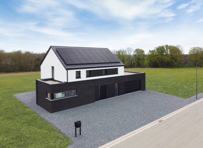 Maison moderne à deux étages avec un extérieur noir et blanc, de grands panneaux solaires sur le toit, une allée en gravier et entourée d'herbe verte et d'arbres sous un ciel partiellement nuageux.