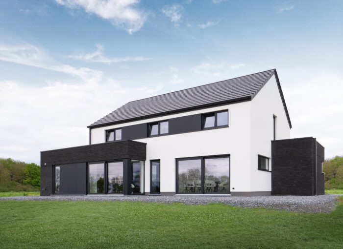 Maison moderne à deux étages avec un extérieur blanc et noir, de grandes fenêtres et un toit en pente, située sur une cour herbeuse sous un ciel partiellement nuageux.