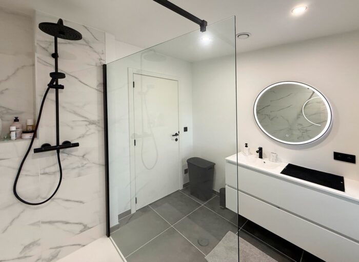 Salle de bain moderne avec douche à l'italienne dotée d'une pomme de douche à effet pluie noire, d'un mur à motifs de marbre, d'un séparateur en verre, d'une vanité blanche avec un miroir rond rétroéclairé et d'un sol carrelé gris.