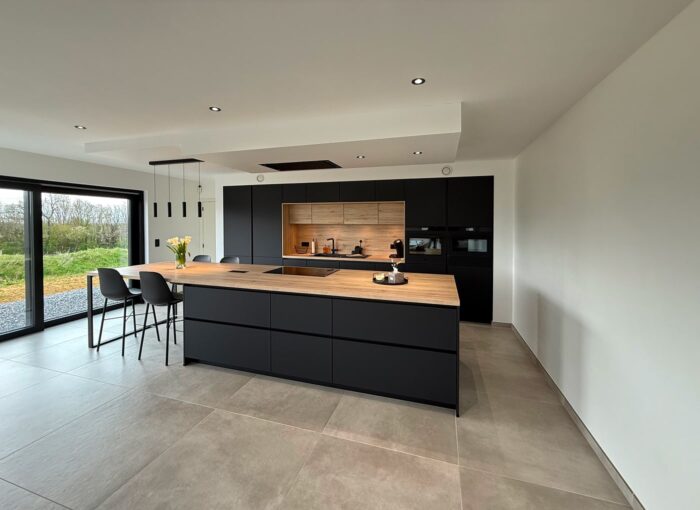 Cuisine moderne avec placards noir mat, îlot central en bois, électroménagers encastrés et tabourets de bar. De grandes fenêtres laissent entrer la lumière naturelle et une décoration minimaliste crée une atmosphère épurée et ouverte.