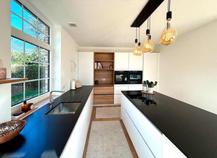 Cuisine moderne avec armoires blanches, plans de travail noirs, suspensions, four encastré, grande fenêtre laissant entrer la lumière naturelle et décoration minimaliste. Un tapis est placé entre les deux plans de travail.