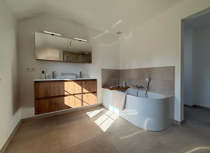 Salle de bains moderne avec meuble-lavabo en bois, grand miroir, double vasque et baignoire ronde blanche dans un angle. Le sol carrelé beige baigne la pièce de lumière, créant une atmosphère lumineuse et aérée.