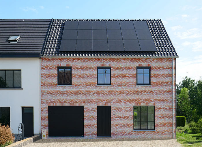 Een modern bakstenen huis met zwarte kozijnen en een garagedeur, met zonnepanelen op het donkere pannendak. Bomen en groen zijn zichtbaar op de achtergrond onder een strakblauwe lucht.