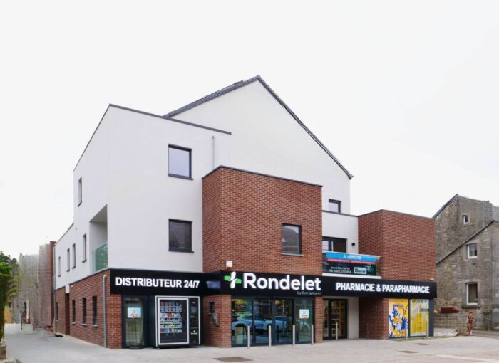 Une pharmacie moderne aux façades blanches et aux briques rouges, avec de grandes fenêtres et une enseigne « Pharmacie & Parapharmacie Rondelet ». Un distributeur automatique et une fresque murale sont présents près de l'entrée.