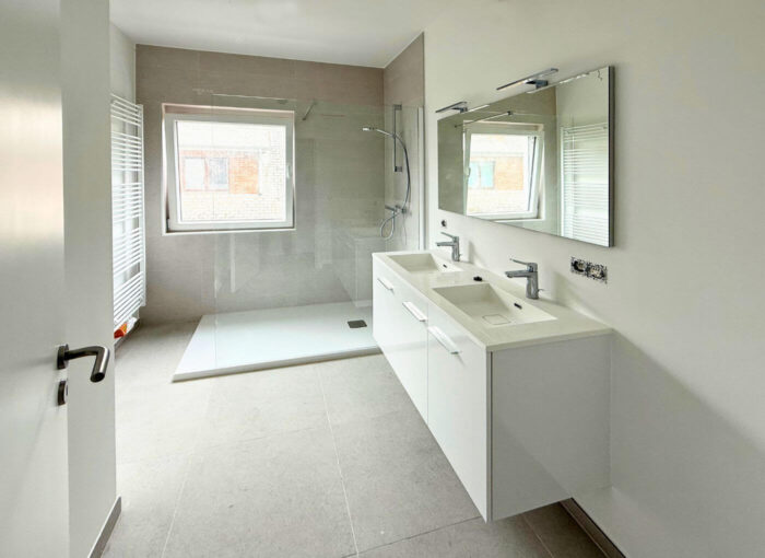 Salle de bain moderne avec douche à l'italienne, grande fenêtre, double vasque avec plan de travail blanc, deux miroirs et robinetterie minimaliste. L'espace est lumineux grâce à son carrelage et ses murs aux tons neutres.