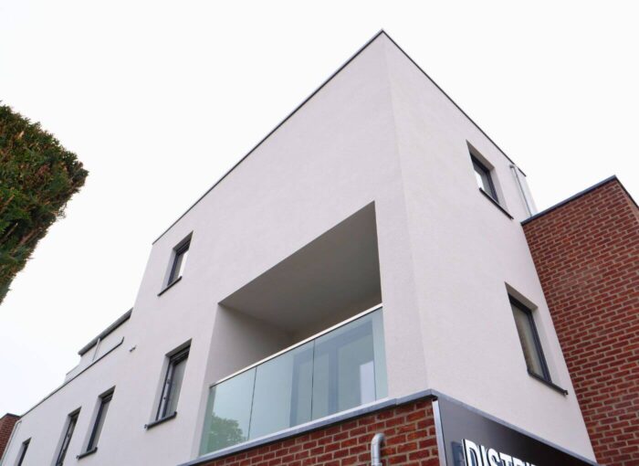 Un bâtiment moderne avec une façade blanche, des formes rectangulaires, de grandes fenêtres et un balcon en verre, à côté d'une section de mur de briques rouges ; photo prise sous un angle bas contre un ciel blanc.