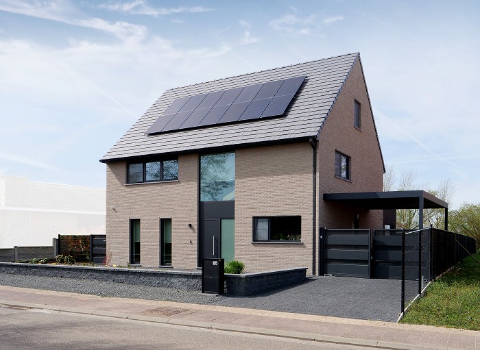 Maison moderne à deux étages avec extérieur en briques beiges, grandes fenêtres, panneaux solaires sur le toit en pente et un abri d'auto couvert sur le côté droit, entourée d'une allée pavée et d'un aménagement paysager minimal.