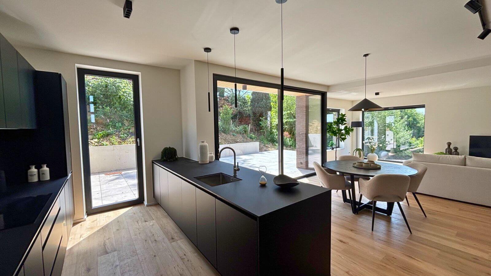 Cuisine ouverte moderne et espace de vie avec îlot noir, table à manger, sols en bois clair, grandes fenêtres et portes s'ouvrant sur un patio ensoleillé avec de la verdure à l'extérieur. La lumière naturelle remplit cet espace élégant et minimaliste.