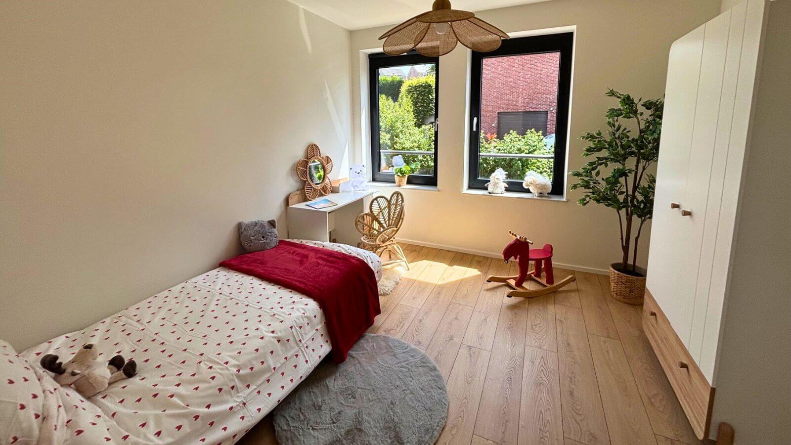 Une chambre d'enfant confortable et lumineuse avec un lit simple, une couverture rouge et une couette à motifs en forme de cœur. Un petit bureau, une chaise en osier, un cheval à bascule, une armoire et une plante décorent l'espace. Deux grandes fenêtres laissent entrer la lumière naturelle.