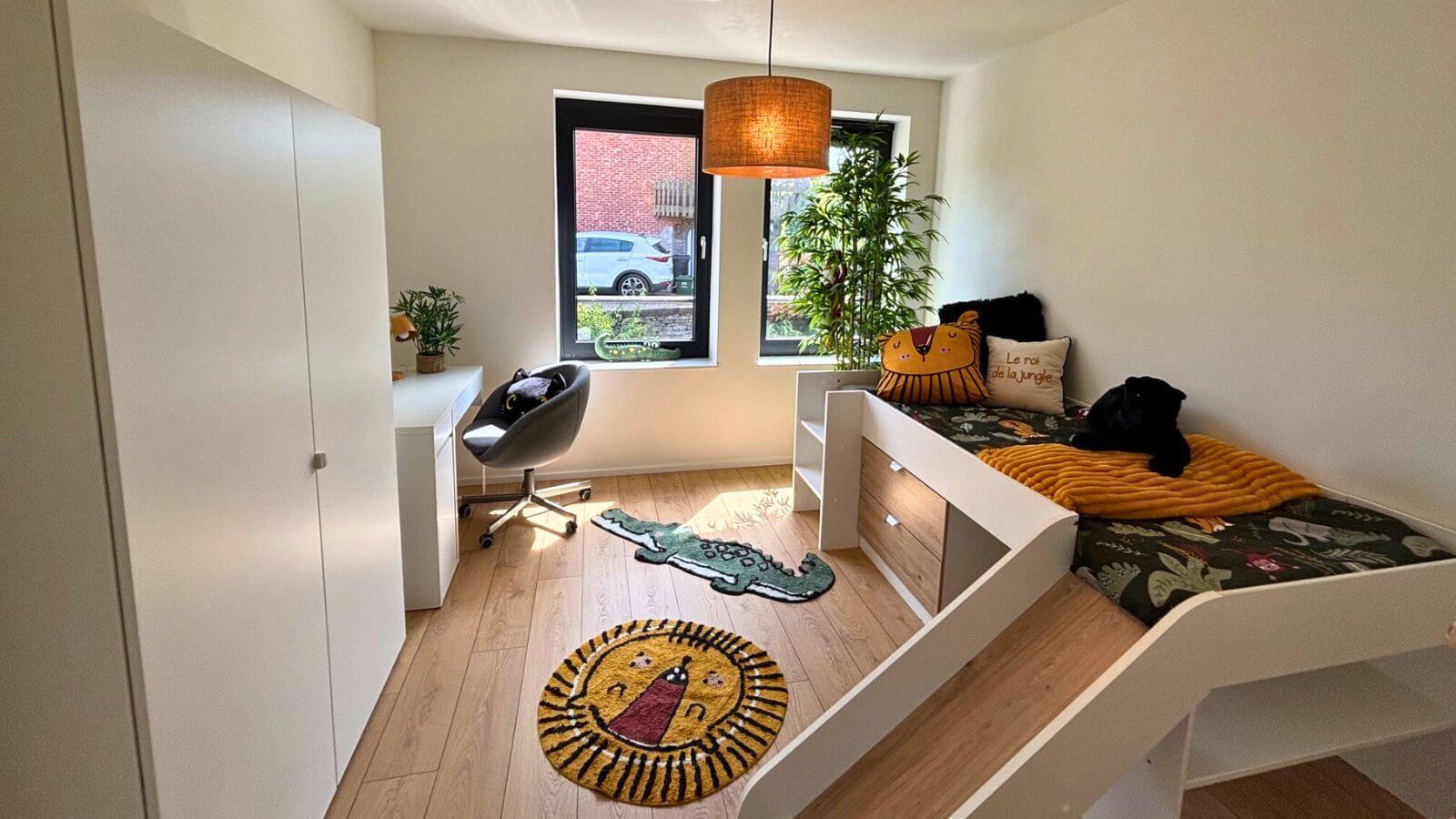 Une chambre d'enfant claire et bien rangée avec un lit équipé d'un toboggan, un tapis sur le thème du lion, un bureau avec une chaise près de la fenêtre et des plantes sur le rebord de la fenêtre. Un animal en peluche noir est posé sur le lit.