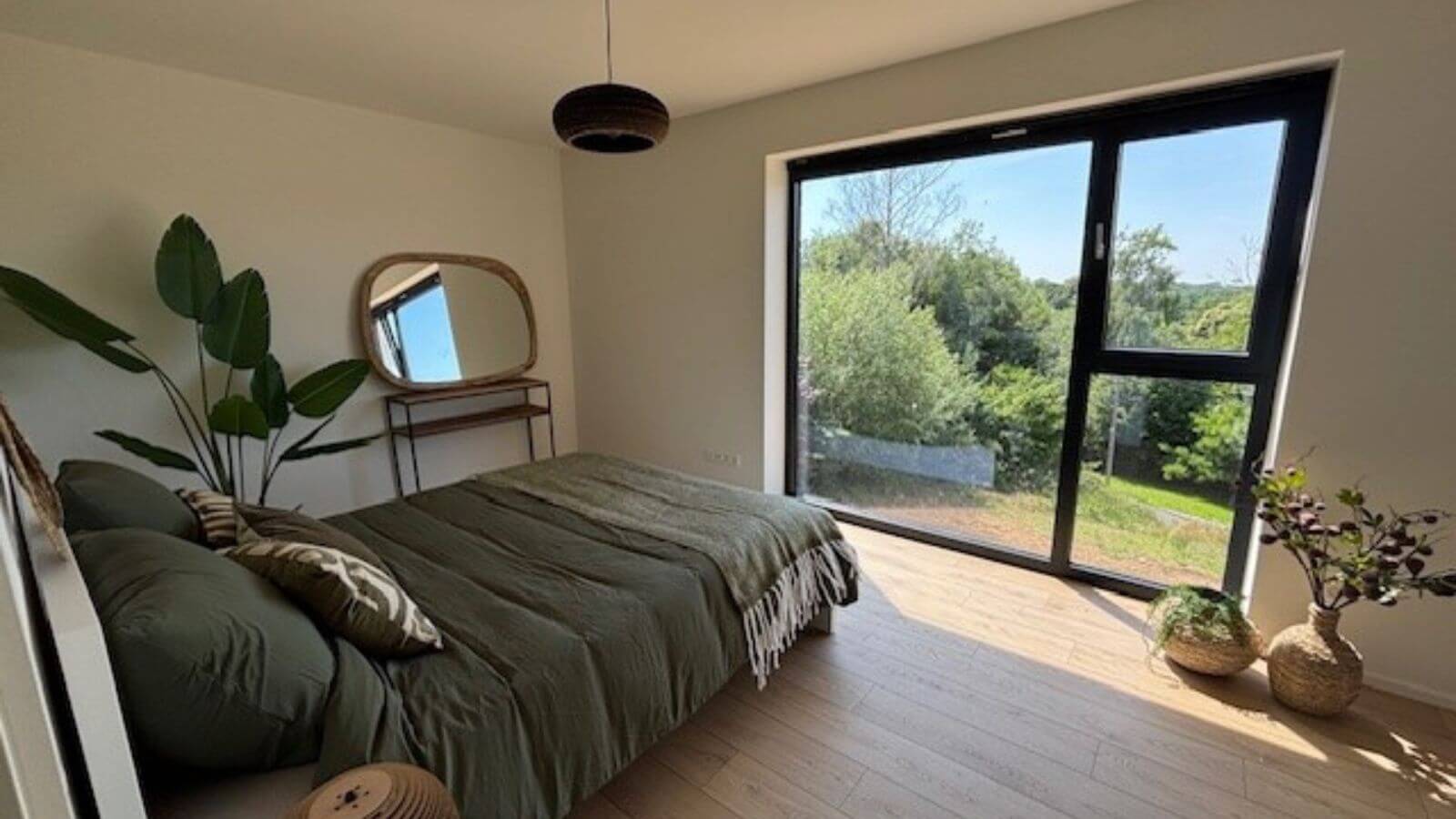 Une chambre lumineuse avec une grande fenêtre montrant la verdure à l'extérieur, un lit avec des draps verts, un miroir sur une petite table, des plantes en pot, un sol en bois clair et une lampe suspendue au plafond.