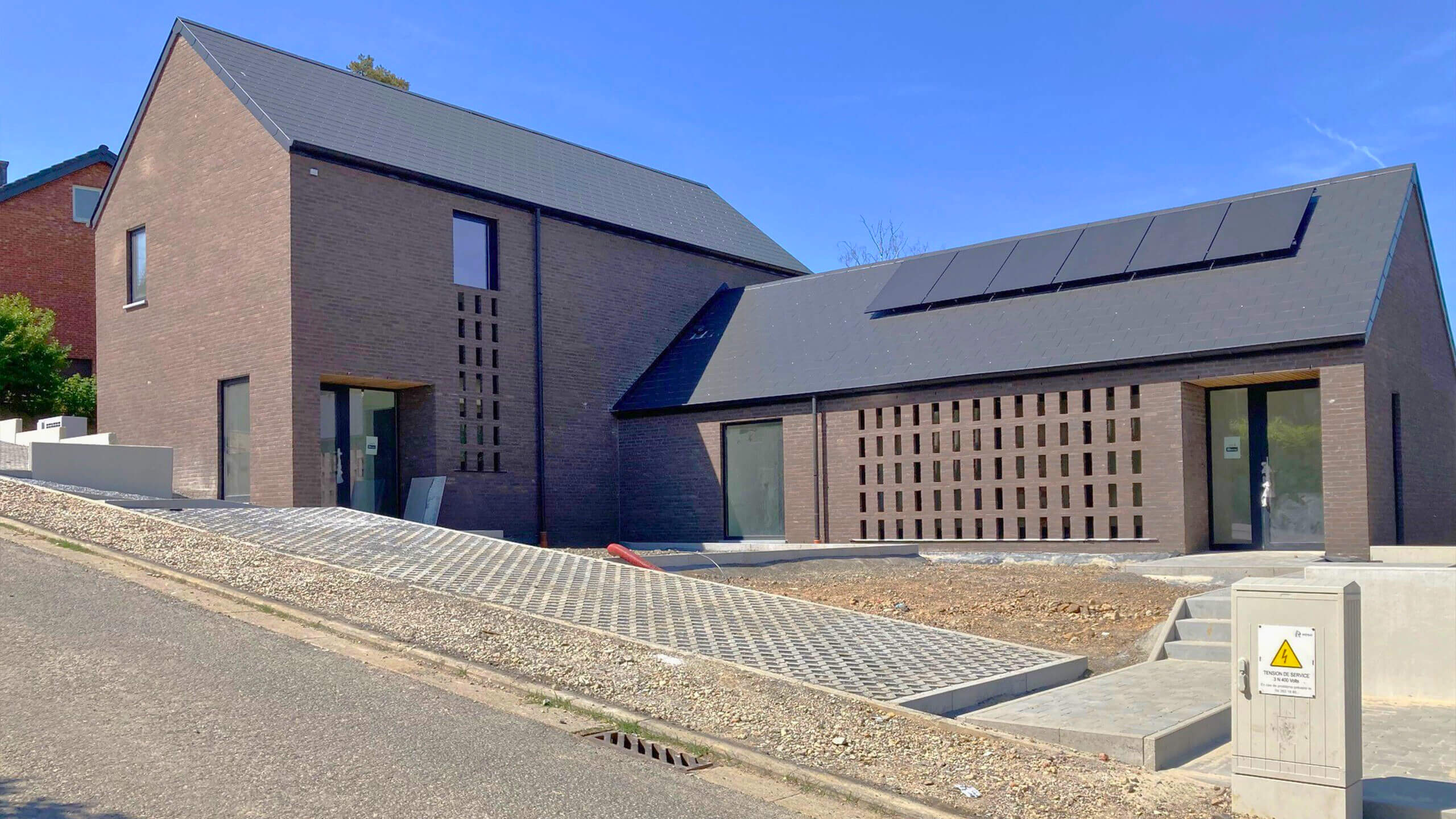 Maison moderne en briques sur une rue en pente, avec fenêtres rectangulaires, panneaux solaires sur le toit et allée pavée. La zone près de l'entrée est en cours de construction, avec terrassement et aménagement paysager inachevé.