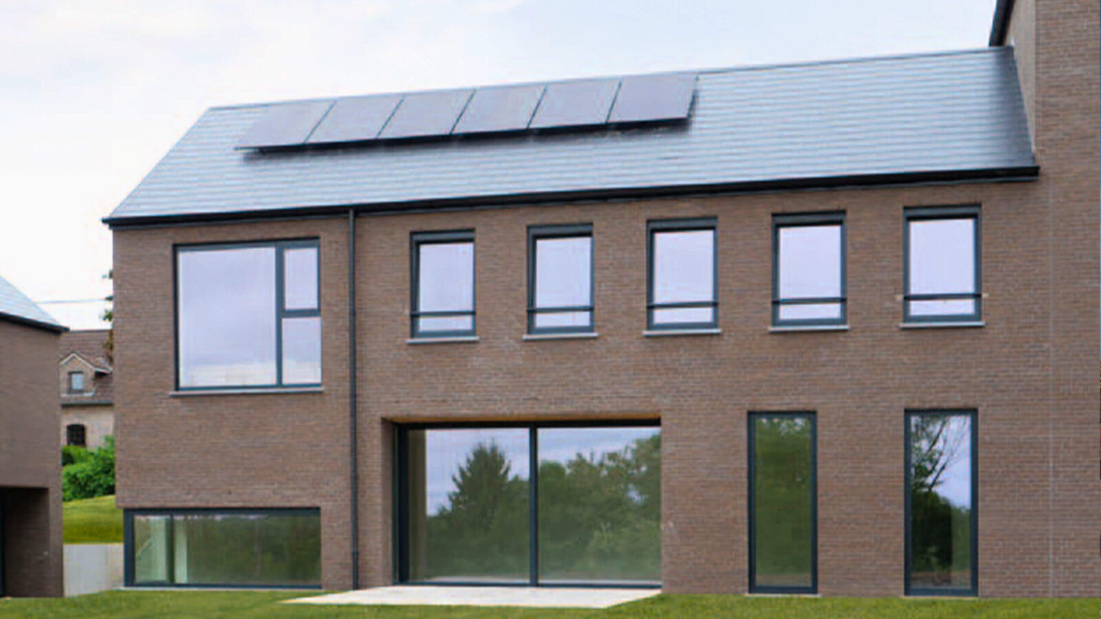 Une maison moderne en briques à deux étages avec de grandes fenêtres et des panneaux solaires installés sur le toit en pente, entourée d'herbe verte et d'arbres.