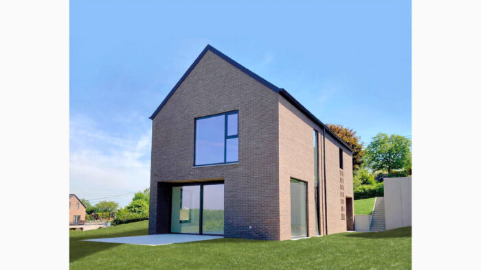 Une maison moderne à deux étages, au toit pentu, dotée de grandes fenêtres et d'une façade en briques, se dresse sur une pelouse verdoyante sous un ciel bleu clair. Des arbres et une autre maison sont visibles en arrière-plan.