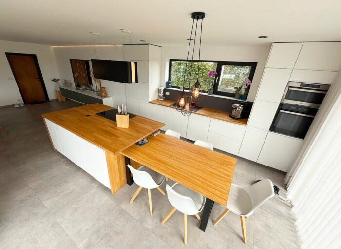 Cuisine moderne avec des armoires blanches, des comptoirs en bois, des appareils encastrés, un grand îlot et une table de salle à manger avec quatre chaises. Des suspensions sont suspendues au-dessus de la table et les fenêtres laissent entrer la lumière naturelle.