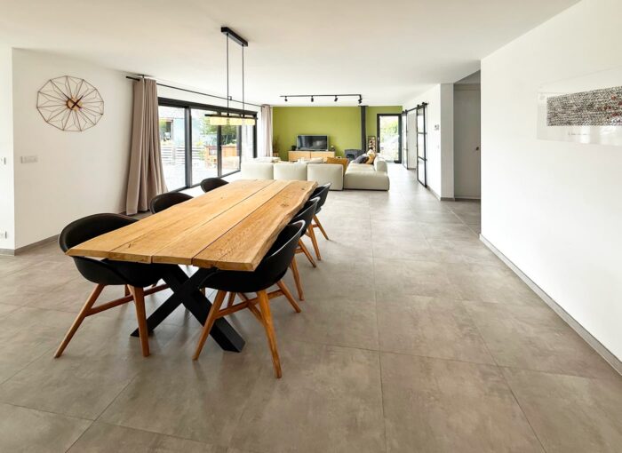Espace de vie ouvert et moderne avec une table de salle à manger en bois, six chaises noires, de grandes dalles de sol grises et un espace de vie avec des canapés blancs en arrière-plan. De grandes fenêtres laissent entrer la lumière naturelle.
