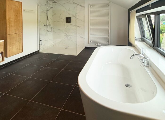 Salle de bains moderne avec une grande baignoire blanche, un sol en carrelage noir, une douche vitrée aux murs de marbre, un radiateur sèche-serviettes mural et de larges fenêtres laissant entrer la lumière naturelle.