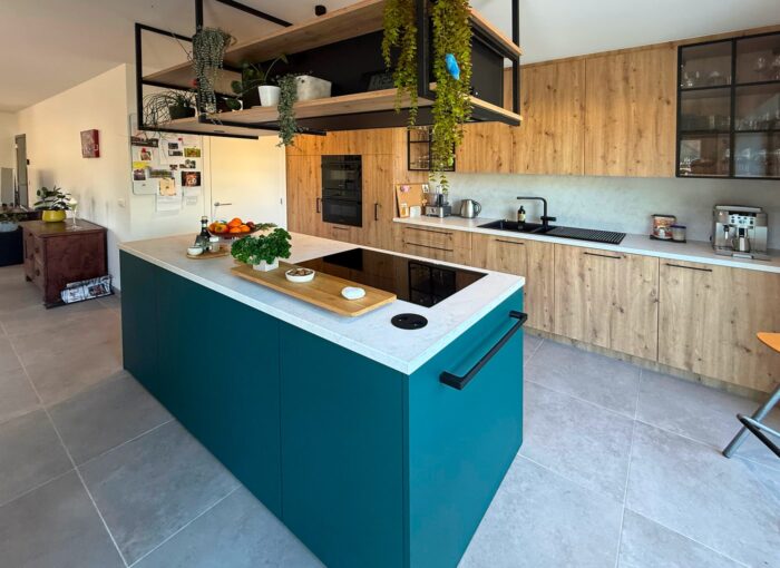 Moderne keuken met een groenblauw eiland, wit aanrechtblad, ingebouwd fornuis, houten kastjes, open planken met planten en verschillende keukenspullen. Lichtgrijze tegelvloer en lichte, netjes georganiseerde ruimte.