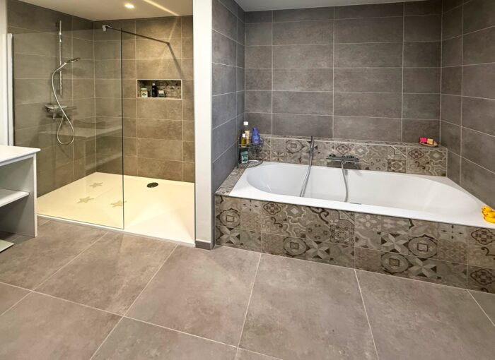 Moderne badkamer met grote grijze tegels, een glazen inloopdouche aan de linkerkant en een rechthoekig bad met tegelrand met patroon aan de rechterkant. Flessen met toiletartikelen zijn zichtbaar bij het bad en in de douche.