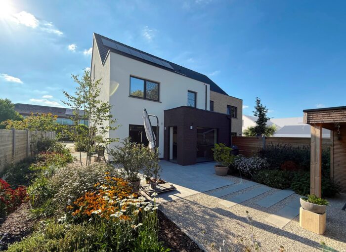 Modern huis van twee verdiepingen met zonnepanelen, een mix van witte en donkere buitenkant, grote ramen en een aangelegde achtertuin met bloemen en bomen onder een strakblauwe hemel.