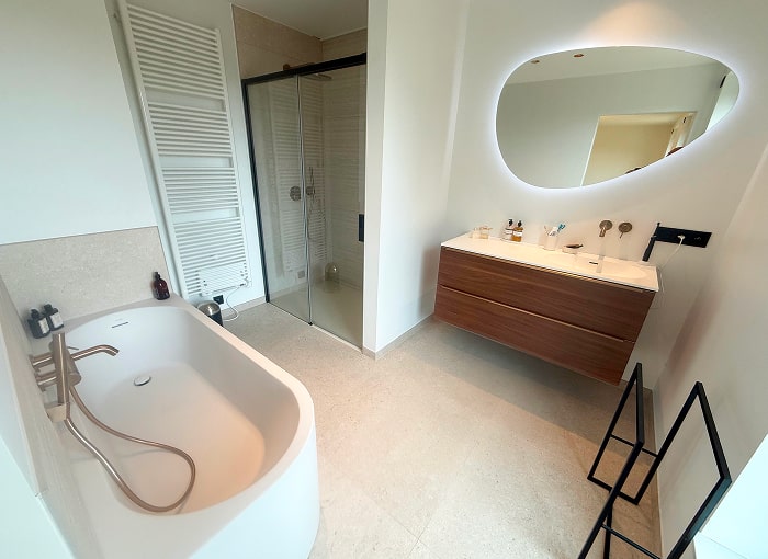 Moderne badkamer met vrijstaand bad, wandkraan, glazen douchewand, zwevend houten badmeubel met wastafel, grote ovale spiegel met achtergrondverlichting en handdoekradiator. Minimalistisch design met lichtgekleurde vloeren.