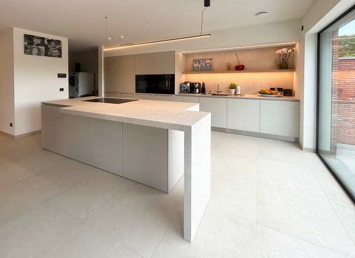 Moderne, minimalistische keuken met lichtgrijze kasten en een groot eiland, ingebouwde apparatuur, verlichting onder de kasten en grote ramen die natuurlijk licht binnenlaten. Decoratieve items en kleine apparaten staan op het aanrecht achteraan.