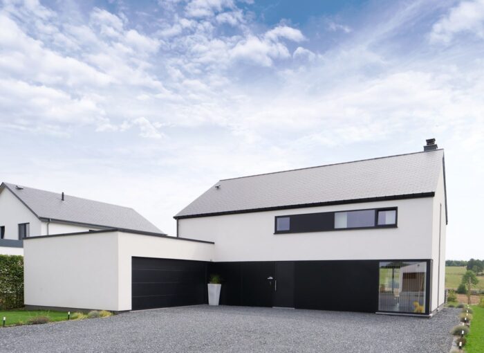 Een modern huis van twee verdiepingen met een plat en schuin dak, met zwart-witte buitenmuren, grote ramen, een garage en een oprit van grind onder een gedeeltelijk bewolkte hemel.