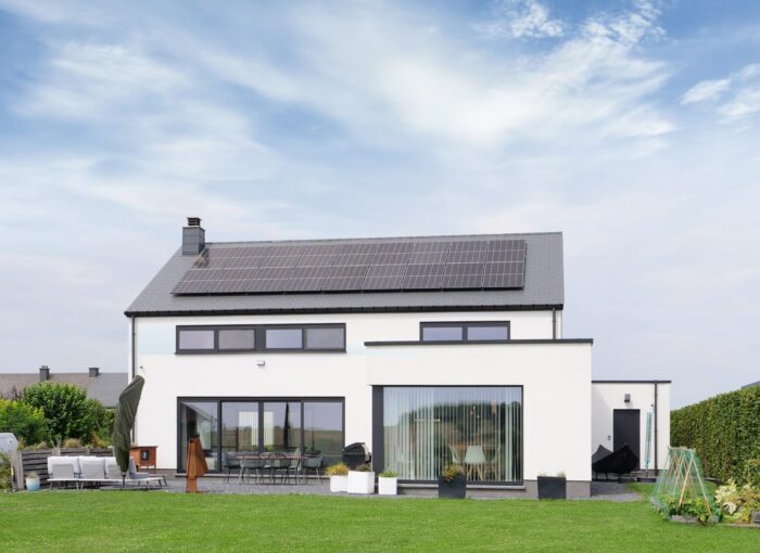 Une maison moderne à deux étages avec de grandes fenêtres, des murs blancs et des panneaux solaires sur le toit est posée sur une pelouse verte sous un ciel partiellement nuageux. Des meubles d'extérieur et un petit jeu sont visibles dans la cour.