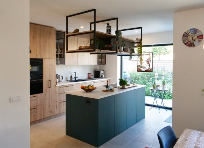 Moderne keuken met houten kastjes, groenblauw eiland, hangende planken met planten en een groot raam dat uitkijkt op een kleine patio met tafel en stoelen. Aan de rechtermuur hangt een kleurrijke wandklok.