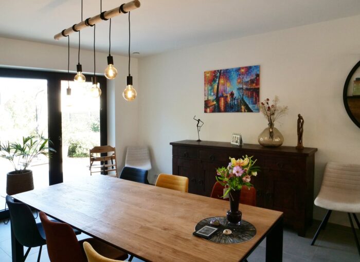Een moderne eetkamer met een houten tafel, kleurrijke stoelen en hangende lampen. Er staat een boeket op tafel en er is een dressoir met decor, kunst aan de muur en grote ramen die natuurlijk licht binnenlaten.
