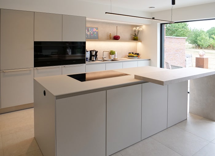 Moderne keuken met strakke, minimalistische witte kasten, een ingebouwde oven en een centraal eiland met een inductiekookplaat. Een groot raam kijkt uit op een groene buitenruimte, waardoor er natuurlijk licht in de ruimte valt.