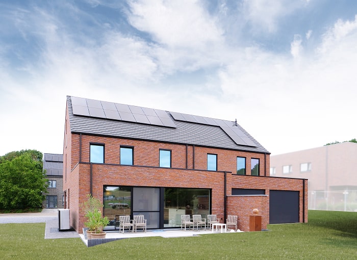 Een modern bakstenen huis van twee verdiepingen met zonnepanelen op het dak, grote ramen, een patio met buitenmeubilair, planten in potten en een groen gazon onder een gedeeltelijk bewolkte hemel.