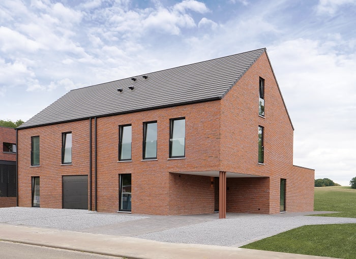 Modern huis van twee verdiepingen met rode bakstenen buitenkant, hoge smalle ramen en een donkergrijs zadeldak. De oprit en de omgeving zijn bedekt met grind en het huis staat tegen een grasveld onder een gedeeltelijk bewolkte hemel.
