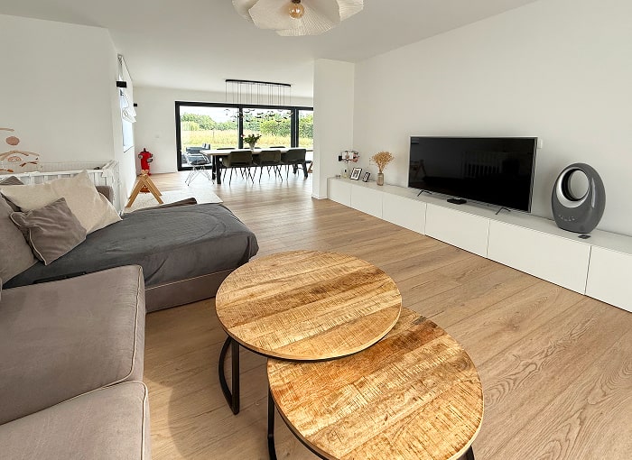 Salon moderne avec un sol en bois clair, un canapé sectionnel gris, deux tables basses rondes en bois, une télévision à écran plat sur un meuble multimédia blanc, et de grandes fenêtres donnant sur une salle à manger avec vue sur la verdure.