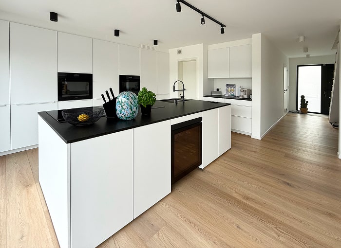 Cuisine moderne avec des armoires blanches, un îlot au comptoir noir avec un évier intégré, un refroidisseur de vin et des objets de décoration. Revêtement de sol en bois clair et design minimaliste. Le couloir mène à une porte à l'arrière-plan.