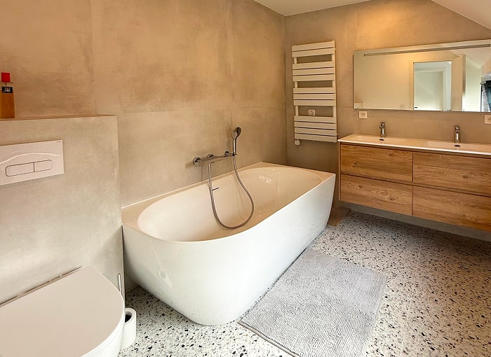 Salle de bains moderne avec baignoire blanche autoportante, robinetterie murale, meuble-lavabo à double vasque en bois avec grand miroir, porte-serviettes chauffant, toilettes, murs beiges et sol en terrazzo.